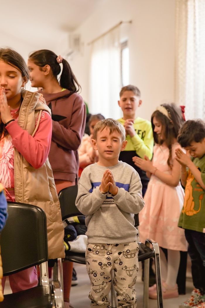 Children gathered in prayer with hands clasped at an indoor event.